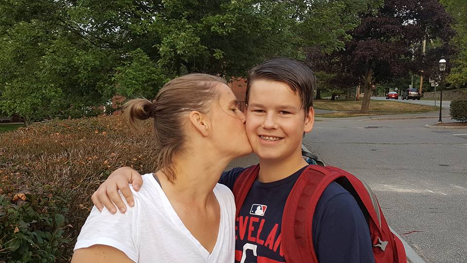 a young teenage boy wearing a backback getting kissed by his mom